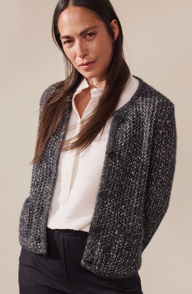 Eine Frau trägt eine schlichte, weiße Bluse, kombiniert mit heller oder dunkler Hose. In einem Look ergänzt sie das Outfit mit einer dunklen Strickjacke für einen klassisch-eleganten Stil.