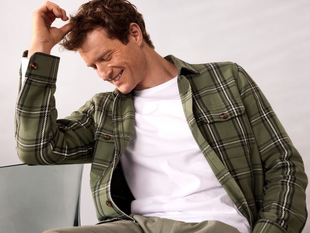 Ein Mann sitzt auf einem Stuhl, schaut nach unten zur Seite und trägt ein Karo Overshirt in Schilf, darunter ein weißes Shirt. Ein Mann sitzt auf einem Stuhl, schaut nach unten zur Seite und trägt ein Karo Overshirt in Schilf, darunter ein weißes Shirt.