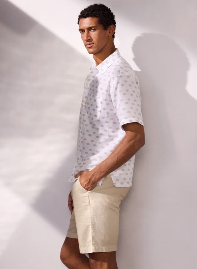 Ein Mann trägt ein weiß-bedrucktes Leinen-Mix Revers-Kragen-Hemd und hält die Hände in den Taschen einer Leinenmix Bermudas. Ein Mann trägt ein weiß-bedrucktes Leinen-Mix Revers-Kragen-Hemd und hält die Hände in den Taschen einer Leinenmix Bermudas.