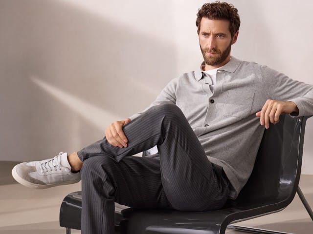 Ein Mann sitzt entspannt auf einem modernen Stuhl und trägt ein geknöpftes Strickpolo in Grau, dazu die gestreifte Macht-Alles-Mit Hose 2.0. Ein Mann sitzt entspannt auf einem modernen Stuhl und trägt ein geknöpftes Strickpolo in Grau, dazu die gestreifte Macht-Alles-Mit Hose 2.0.