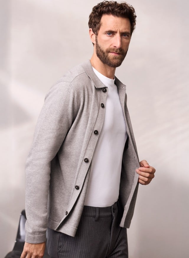 Ein Mann trägt ein geknöpftes Strickpolo in Grau, darunter ein weißes Shirt und dazu eine dunkele Hose. Ein Mann trägt ein geknöpftes Strickpolo in Grau, darunter ein weißes Shirt und dazu eine dunkele Hose.