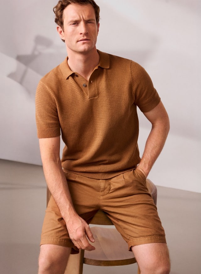 Ein Mann sitzt auf einem Hocker und trägt das Strickpolo Klimastruktur in der Farbe Latte, dazu eine Bermudas. Ein Mann sitzt auf einem Hocker und trägt das Strickpolo Klimastruktur in der Farbe Latte, dazu eine Bermudas.