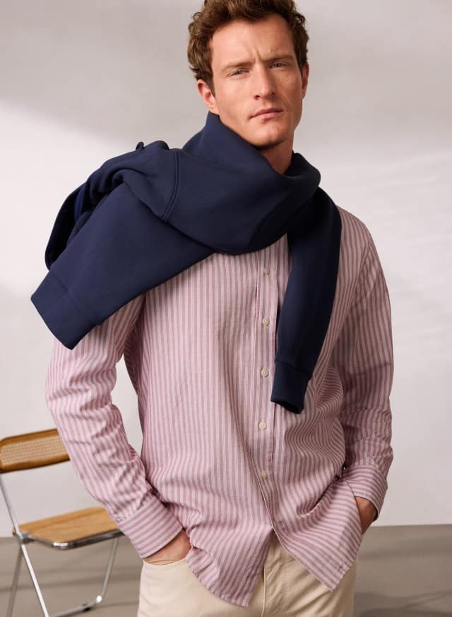 Ein Mann trägt Hemd aus Royal-Oxford mit roten Streifen und hat eine Hoodie quer über die Schultern gebunden. Ein Mann trägt Hemd aus Royal-Oxford mit roten Streifen und hat eine Hoodie quer über die Schultern gebunden.