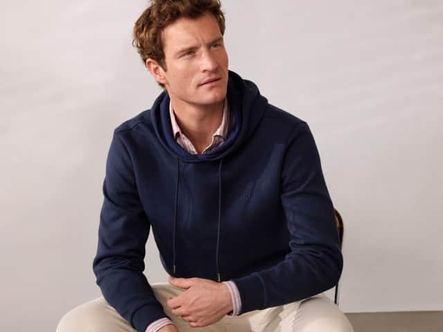 Ein Mann trägt einen Hoodie in Marine, darunter ein rot-gestreiftes Hemd und dazu eine helle Hose. Ein Mann trägt einen Hoodie in Marine, darunter ein rot-gestreiftes Hemd und dazu eine helle Hose.