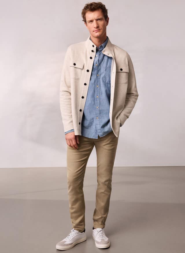 Ein Mann trägt eine helle Sweatjacke mit Stehkragen, darunter ein blaues Hemd und dazu eine helle Hose. Ein Mann trägt eine helle Sweatjacke mit Stehkragen, darunter ein blaues Hemd und dazu eine helle Hose.