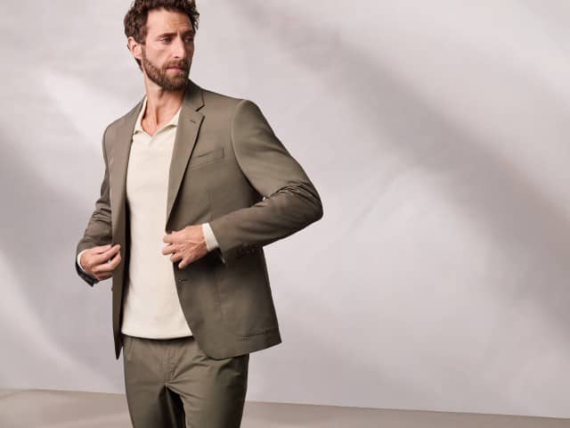 Ein Mann trägt ein Anzug aus Baumwollmix in Khaki, darunter einen gerippten Polopullover in Natur. Ein Mann trägt ein Anzug aus Baumwollmix in Khaki, darunter einen gerippten Polopullover in Natur.