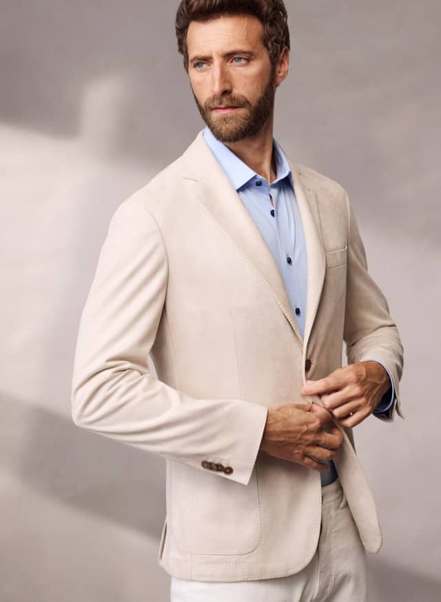Ein Mann trägt ein Sakko aus Waschvelours in Beige, darunter ein hellblaues Hemd und dazu eine helle Hose. Ein Mann trägt ein Sakko aus Waschvelours in Beige, darunter ein hellblaues Hemd und dazu eine helle Hose.