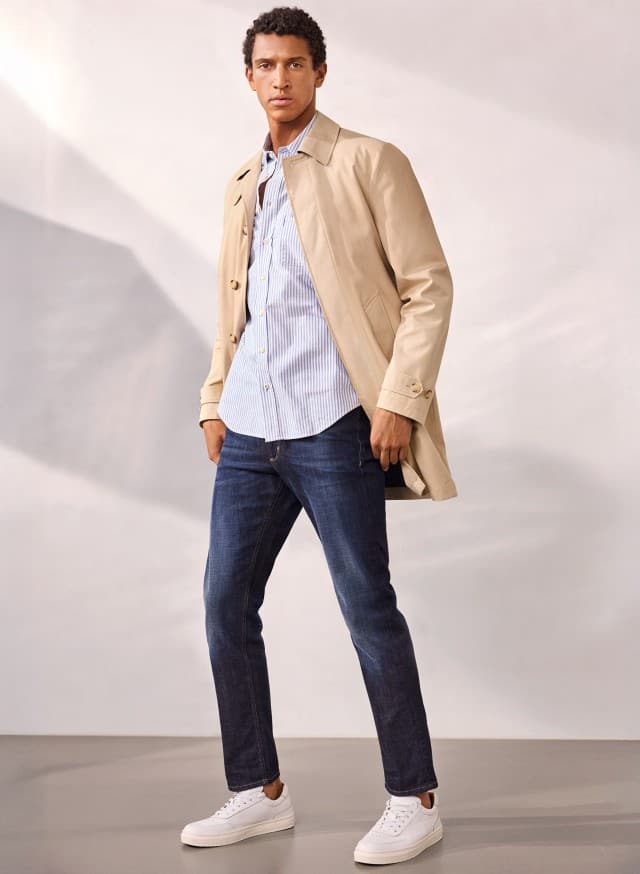 Ein Mann trägt einen Baumwoll Mantel 2 in 1 in Beige, darunter ein Hemd in hellblau, dazu eine Jeans und weiße Sneaker. Ein Mann trägt einen Baumwoll Mantel 2 in 1 in Beige, darunter ein Hemd in hellblau, dazu eine Jeans und weiße Sneaker.