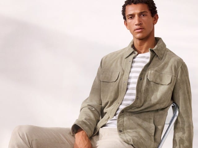 Ein Mann trägt sitzt lässig auf einem Stuhl und eine Velourslederhemdjacke in der Farbe Salbei, darunter ein gestreiftes Shirt und dazu eine helle Hose. Ein Mann trägt sitzt lässig auf einem Stuhl und eine Velourslederhemdjacke in der Farbe Salbei, darunter ein gestreiftes Shirt und dazu eine helle Hose.