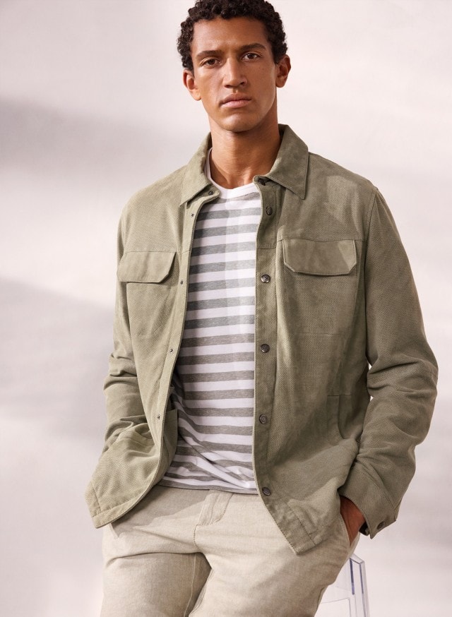 Ein Mann trägt eine Velourslederhemdjacke in der Farbe Salbei, darunter ein gestreiftes Shirt und dazu eine helle Hose. Ein Mann trägt eine Velourslederhemdjacke in der Farbe Salbei, darunter ein gestreiftes Shirt und dazu eine helle Hose.