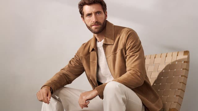 Ein Mann sitzt auf einem Sessel und trägt einen Ziegenvelours Blouson in Beige, darunter einen hellen Pullover und dazu eine helle Hose. Ein Mann sitzt auf einem Sessel und trägt einen Ziegenvelours Blouson in Beige, darunter einen hellen Pullover und dazu eine helle Hose.