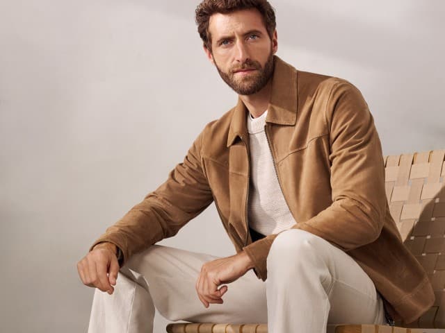 Ein Mann sitzt auf einem Sessel und trägt einen Ziegenvelours Blouson in Beige, darunter einen hellen Pullover und dazu eine helle Hose. Ein Mann sitzt auf einem Sessel und trägt einen Ziegenvelours Blouson in Beige, darunter einen hellen Pullover und dazu eine helle Hose.