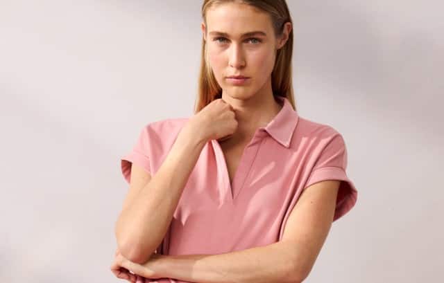 Eine Person trägt ein rosafarbenes kurzärmliges Oberteil mit Kragen und passender Hose. Sie hält einen Arm vor dem Körper verschränkt und berührt mit der anderen Hand ihr Kinn.