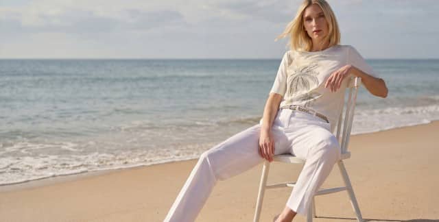 Frau in heller Sommerkleidung mit weißer Hose und T-Shirt mit Palmenmotiv sitzt lässig auf einem Stuhl am Strand. Der Stil wirkt entspannt und elegant.
