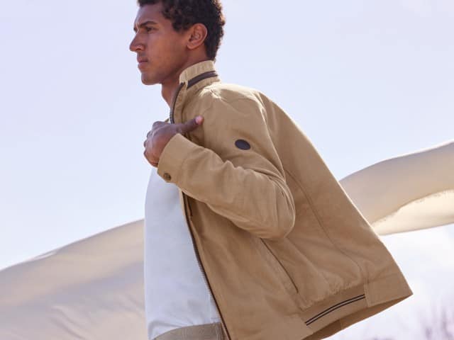 Ein Mann trägt den Leinenblouson Leicht und Luftig in der Farbe Latte, darunter ein weißes Shirt, hinter ihm weht ein großes Leinentuch.