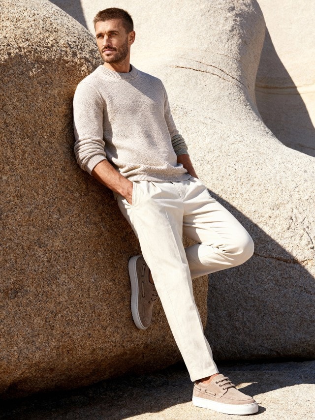 Ein Mann an einen Felsen gelehnt, trägt einen Premium Leinen-Pullover in Natur, dazu eine passende helle Hose.