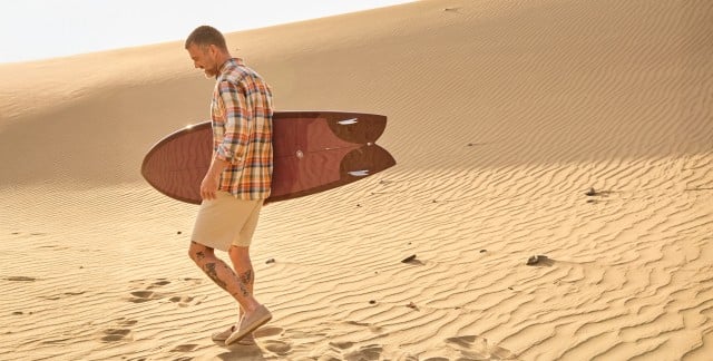 Ein Mann in der Wüste mit einem Surfbrett trägt ein kariertes Hemd und Shorts.