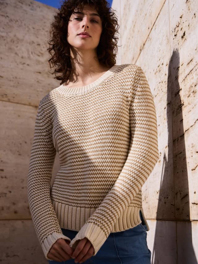 Eine Frau steht vor einer Natursteinwand und trägt einen Pullover mit dezentem Streifen in Creme, dazu eine Marlene Jeans.