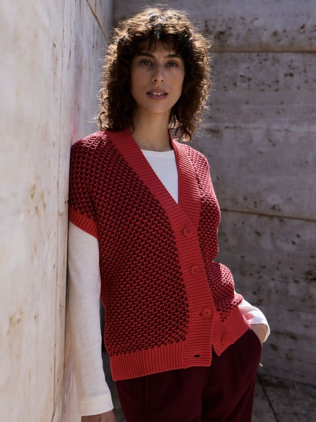 Eine Frau steht vor einer Natursteinwand und trägt eine Strickweste mit Waffelstruktur in Signalrot, darunter ein weißes Langarm-Shirt und dazu eine Jeans.