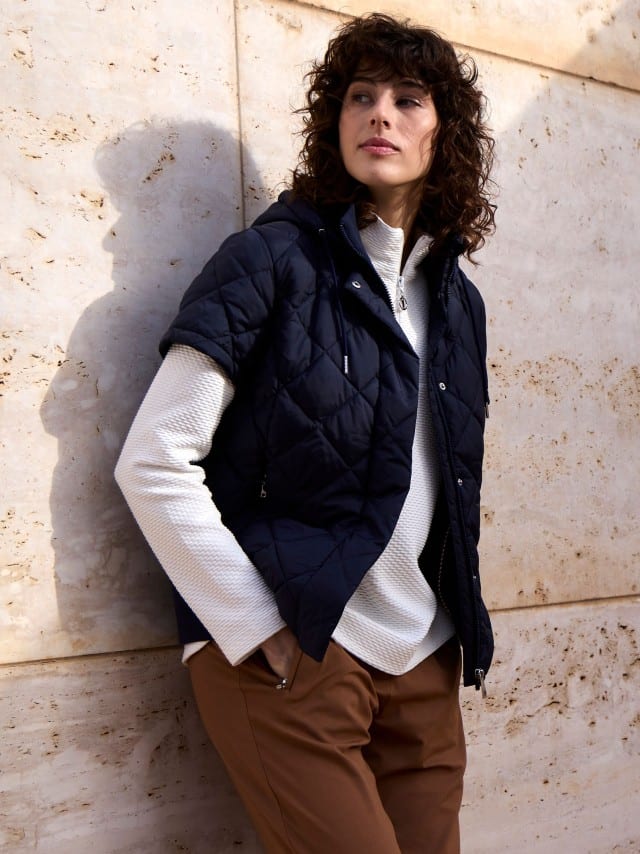 Eine Frau steht an einer Natursteinwand und trägt eine Steppweste Hoodie in Marine, darunter eine Troyer-Sweatshirt in Weiß und dazu eine Hose in Toffee.