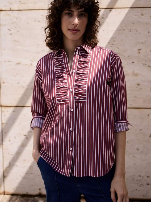 Ein Frau vor einer Natursteinwand trägt eine gestreifte Hemdbluse in Chianti.