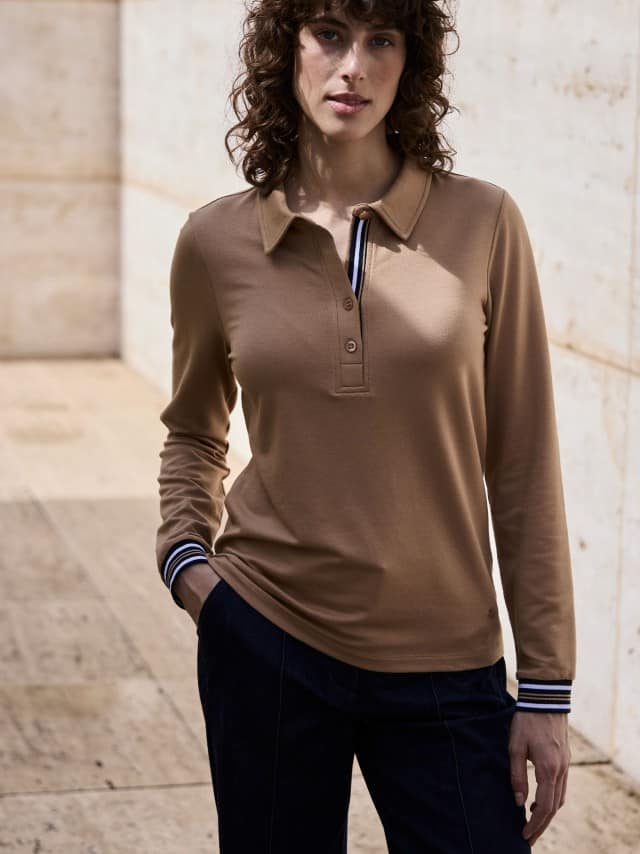 Eine Frau steht vor einer Natursteinwand und trägt ein Poloshirt Kontrastdetail in der Farbe Praline, dazu eine Marlene-Jeans.