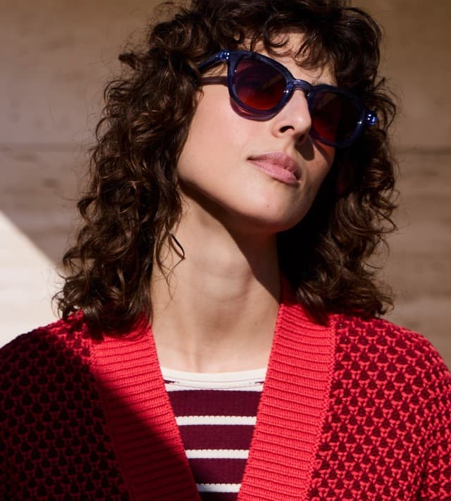 Eine Frau mit lockigem Haar und Sonnenbrille, trägt eine Strickweste mit Waffelstruktur in Signalrot, darunter ein passendes gestreiftes Shirt.