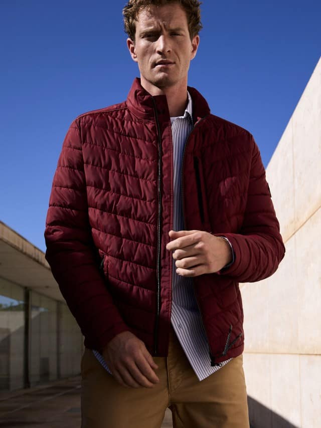 Ein Mann trägt die rote Klima Leichtsteppjacke, darunter ein Hemd in Hellblau und dazu eine Chino in Beige. Ein Mann trägt die rote Klima Leichtsteppjacke, darunter ein Hemd in Hellblau und dazu eine Chino in Beige.