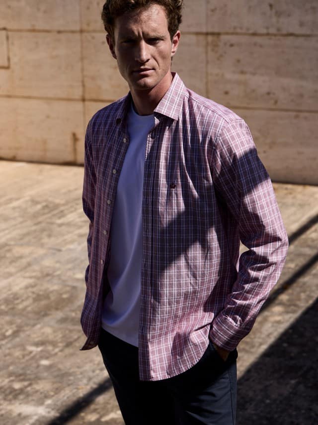 Ein Mann vor einer Natursteinwand trägt das Royal-Oxford Hemd in Karo-Rot, darunter ein weißes Shirt und dazu eine Chino in Beige. Ein Mann vor einer Natursteinwand trägt das Royal-Oxford Hemd in Karo-Rot, darunter ein weißes Shirt und dazu eine Chino in Beige.