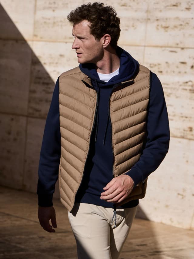 Ein Mann vor einer Natursteinwand trägt einen Hoodie in Marine, darüber eine Klima Leichtsteppweste in der Farbe Karamel, dazu eine helle Hose. Ein Mann vor einer Natursteinwand trägt einen Hoodie in Marine, darüber eine Klima Leichtsteppweste in der Farbe Karamel, dazu eine helle Hose.