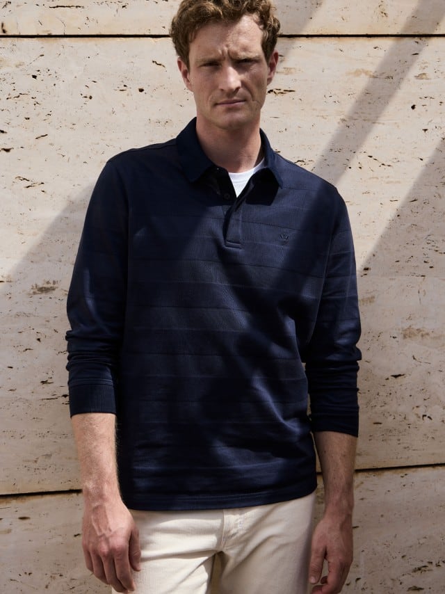 Ein Mann steht vor einer Natursteinwand und trägt das Baumwoll-Polo Webeffekt in Marine, dazu eine helle Jeans. Ein Mann steht vor einer Natursteinwand und trägt das Baumwoll-Polo Webeffekt in Marine, dazu eine helle Jeans.