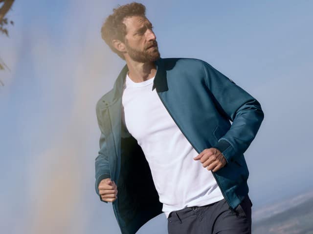 Ein Mann steht auf einem Feld mit gelben Blumen und trägt einen Blouson in Blau, darunter ein weißes Shirt und dazu eine Hose in Marine.