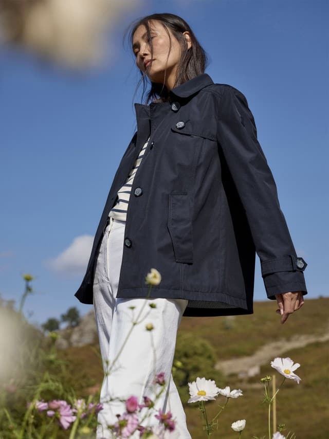Eine Frau steht im Freien vor blauem Himmel und trägt eine dunkelblaue Jacke zu einem gestreiften Oberteil und weißen Hosen.