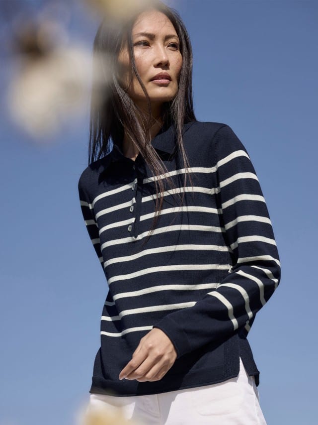 Eine Frau steht im Freien vor blauem Himmel und trägt einen dunkelblauen Pullover mit weißen Streifen zu einer hellen Hose.