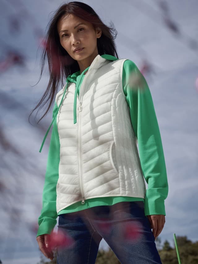 Eine Frau steht im Freien und trägt einen grünen Hoodie unter einer weißen Steppweste sowie eine Jeans.