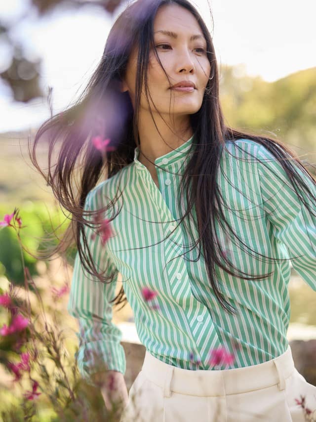 Eine Frau steht im Freien und trägt eine grün‑weiß gestreifte Bluse zu einer hellen Hose.