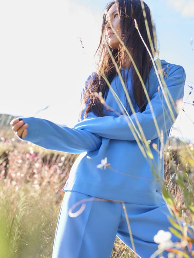 Eine Frau steht in einer Wiese und trägt einen hellblauen Pullover sowie eine farblich passende hellblaue Hose.