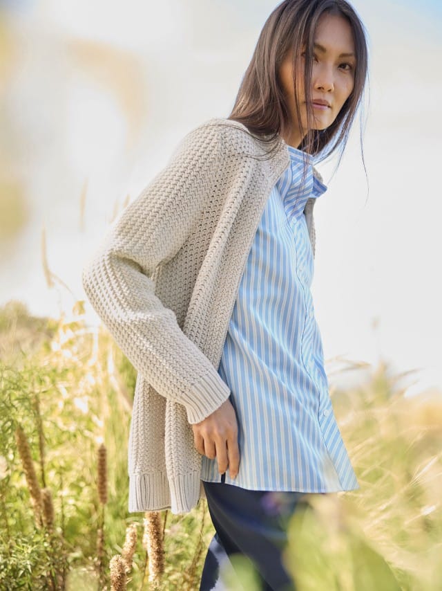 Eine Frau steht im Freien und trägt eine beige Strickjacke über einer hellblau gestreiften Bluse.