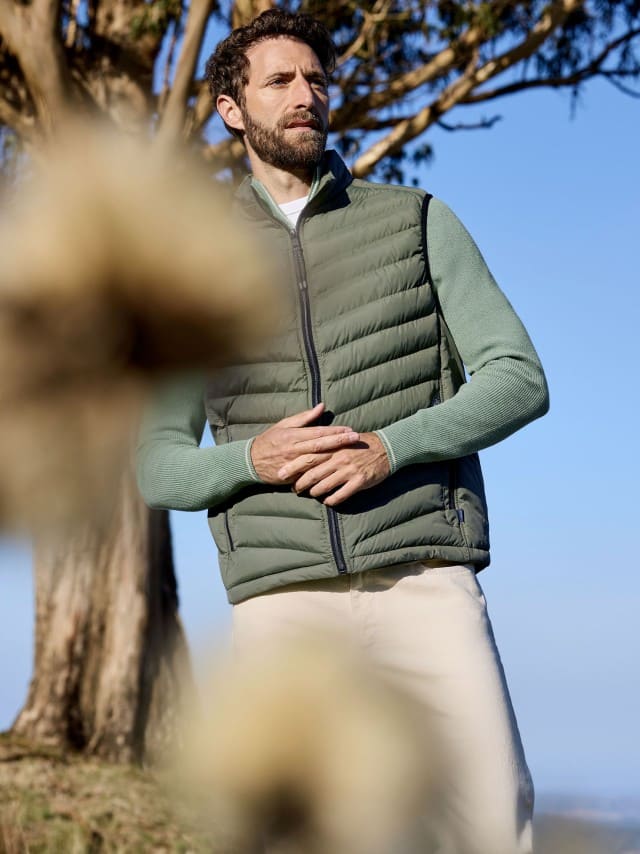 Ein Mann steht vor einem Baum und trägt einen gerippten Baumwoll-Troyer in Pistazie, darüber eine passende Steppweste und dazu eine helle Hose.