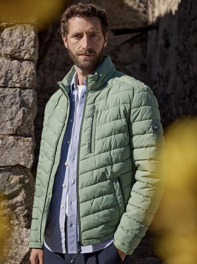 Ein Mann trägt die Klima Leichtsteppjacke in Lindgrün, darunter ein hellblaues Hemd und dazu eine Hose in Marine.