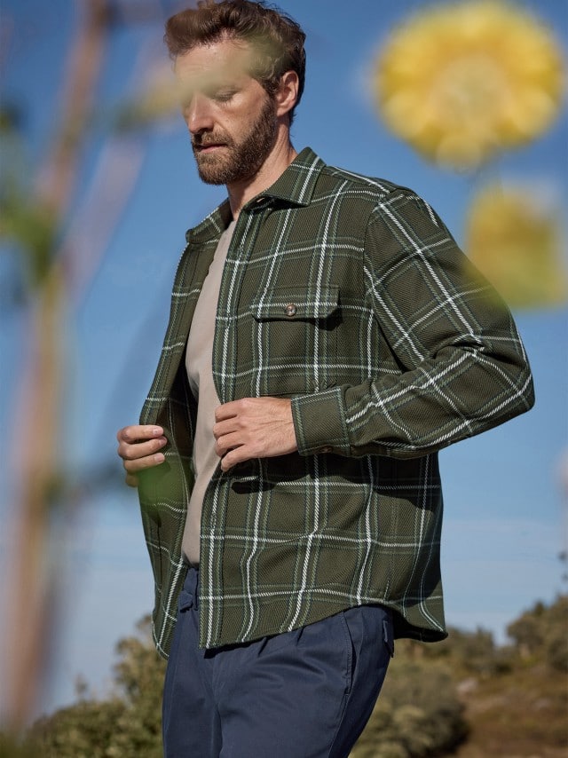 Ein Mann trägt ein Karo Overshirt in Oliv, darunter ein Shirt in Sand und eine Hose in Marine. Im Vordergrund sind gelbe Blüten zu sehen. 
