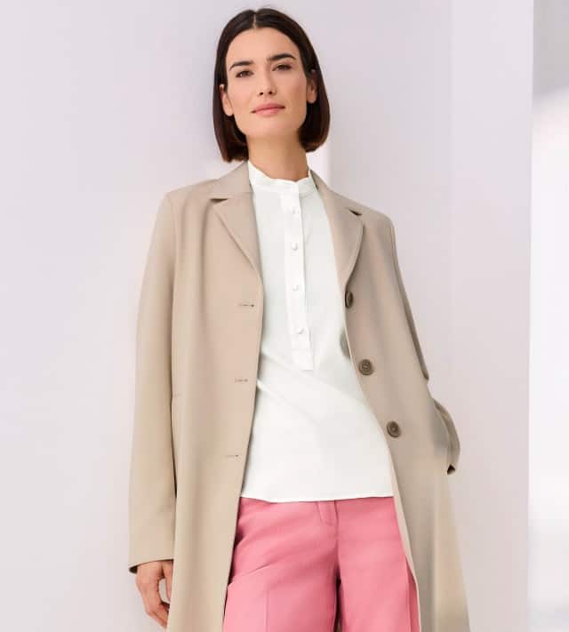 Eine Frau trägt eine beige lange Jacke über einer weißen Bluse und rosa Hose. Der Look wirkt elegant und modern mit einem frischen Farbakzent.