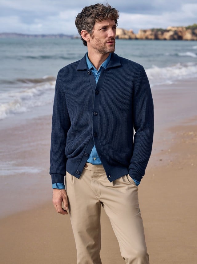 Ein Mann steht am Strand und trägt einen gerippten Polo-Cardigan in Marine, darunter eine Jeans-Hemd und dazu eine Chino in Beige. Ein Mann steht am Strand und trägt einen gerippten Polo-Cardigan in Marine, darunter eine Jeans-Hemd und dazu eine Chino in Beige.