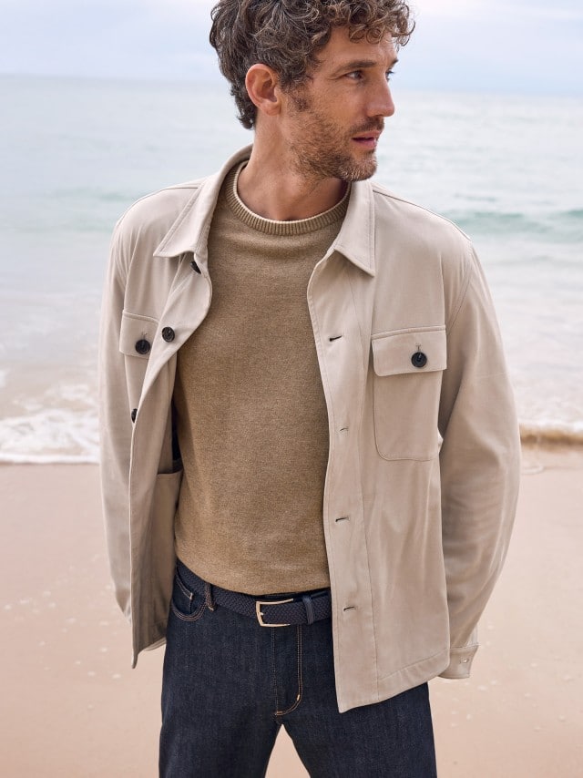 Ein Mann am Strand trägt einen Premium Pullover in Sand Melange, darüber eine Hemdjacke aus Microvelours in Beige und dazu eine Charakter-Jeans. Ein Mann am Strand trägt einen Premium Pullover in Sand Melange, darüber eine Hemdjacke aus Microvelours in Beige und dazu eine Charakter-Jeans.