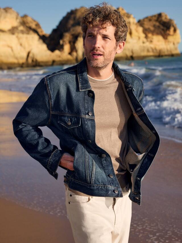 Ein Mann am Strand trägt eine Jeansjacke in Blau, darunter einen Premium Pullover und dazu eine weiße Charakter-Jeans. Ein Mann am Strand trägt eine Jeansjacke in Blau, darunter einen Premium Pullover und dazu eine weiße Charakter-Jeans.