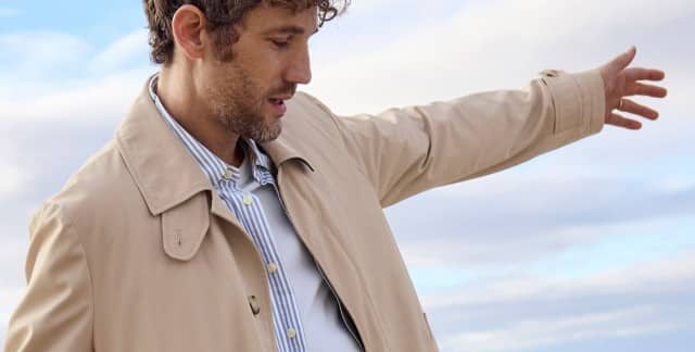 Ein Mann am Strand trägt einen Baumwollmantel in Beige, darunter ein gestreiftes Hemd und ein Shirt. Ein Mann am Strand trägt einen Baumwollmantel in Beige, darunter ein gestreiftes Hemd und ein Shirt.