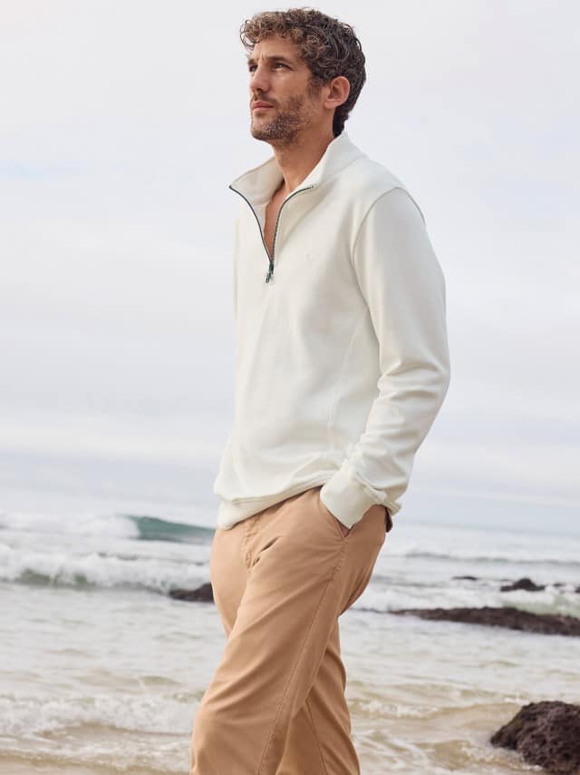 Ein Mann steht am Strand und trägt die 100 Komplimente Chino in Sandgelb, dazu einen Essential Troyer in Offwhite. Ein Mann steht am Strand und trägt die 100 Komplimente Chino in Sandgelb, dazu einen Essential Troyer in Offwhite.