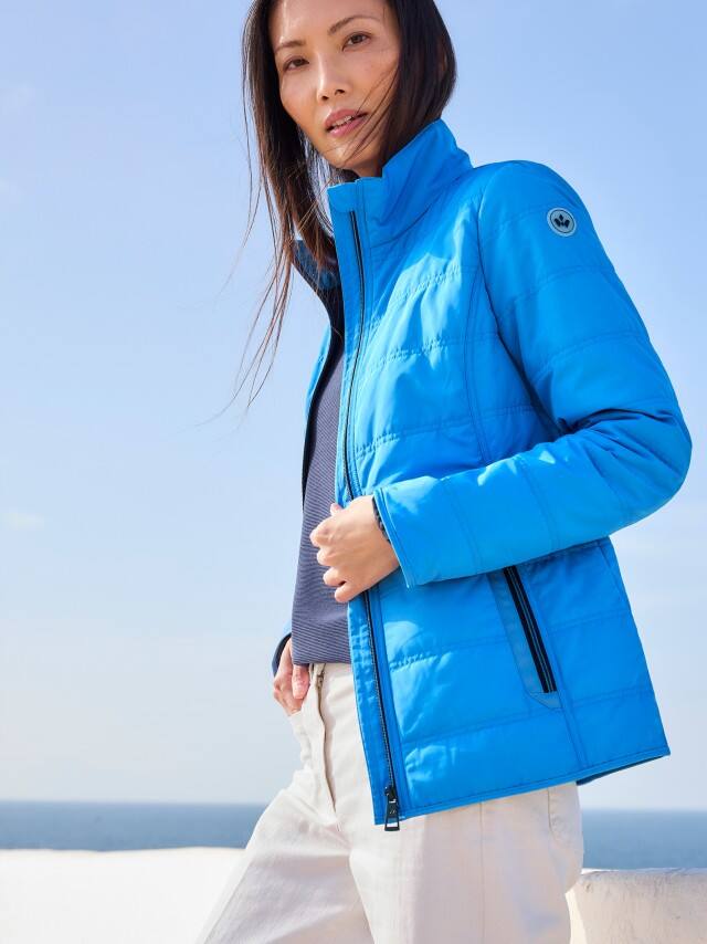 Eine Frau trägt eine leuchtend blaue Steppjacke zu dunkelblauem Shirt und heller Hose, was frisch, sportlich und unkompliziert wirkt. Eine Frau trägt eine leuchtend blaue Steppjacke zu dunkelblauem Shirt und heller Hose, was frisch, sportlich und unkompliziert wirkt.