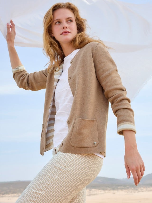 Eine Frau trägt eine Strickjacke in der Farbe Praline, darunter ein weißes Shirt mit Streifenmuster und dazu eine helle Hose.