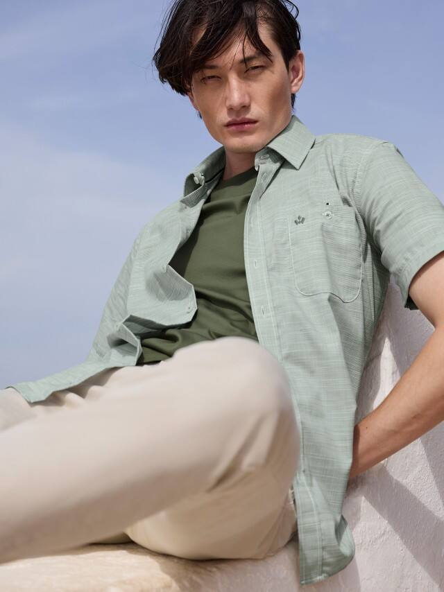 Ein Mann trägt ein hellgrünes Kurzarmhemd über einem olivgrünen Shirt zu beigefarbenen Hosen, was leicht, modern und sommerlich entspannt wirkt.